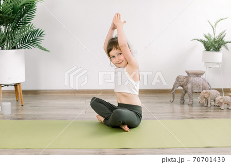 Healthy child girl, doing exercise in sportive top and leggings, practicing yoga at studio indoors. sitting in lotus pose on yoga mat with arms raised up, smiling to camera Healthy child girl, doing exercise in sportive top and leggings, practicing yoga at studio indoors. sitting in lotus pose on yoga mat with arms raised up, smiling to camera 70701439