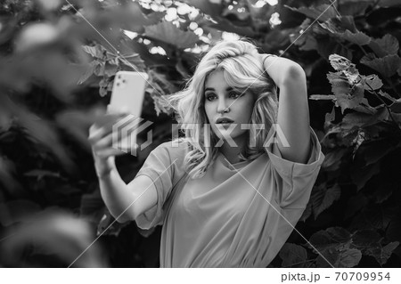 A young beautiful blonde girl is photographed on a smartphone, taking a selfie in the park against of green trees. A young beautiful blonde girl is photographed on a smartphone, taking a selfie in the park against of green trees. 70709954