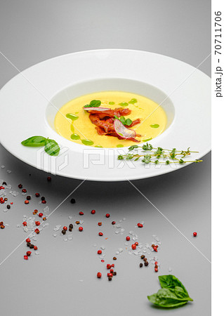 Corn cream soup with smoked meat in a white plate decorated with basil, on a gray background. The first course on the restaurant menu. Corn cream soup with smoked meat in a white plate decorated with basil, on a gray background. The first course on the restaurant menu. 70711706