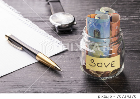 Euro bills and coins in a glass jar with a plate with a blank sheet of paper and a pen and a wrist watch on a black wooden table. Concept of financial savings 70724270