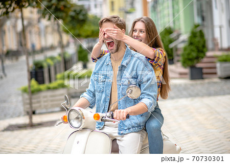 Woman closing her boyfriends eyes while riding a scooter Woman closing her boyfriends eyes while riding a scooter 70730301