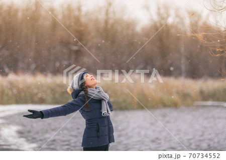 Winter happy girl enjoying snowing snowfall snowflakes falling. Asian lady with open arms wearing cold weather scarf, hat, gloves warm jacket walking outdoor nature woods background 70734552