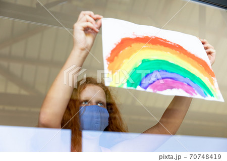 Girl wearing face mask sticking rainbow painting to the window 70748419
