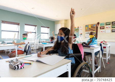 Disable girl wearing face mask raising her hand while sitting on wheelchair in class 70748425