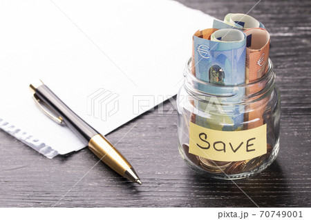 Euro bills and coins in a glass jar with a plate with a blank sheet of paper and a pen on a black wooden table with a sunny reflection on a glossy surface. Savings concept Euro bills and coins in a glass jar with a plate with a blank sheet of paper and a pen on a black wooden table with a sunny reflection on a glossy surface. Savings concept 70749001