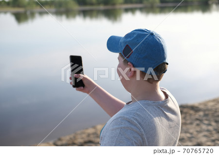 stout teenager boy making selfie. boy standing near the river and looking at smartphone stout teenager boy making selfie. boy standing near the river and looking at smartphone 70765720