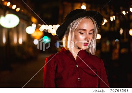 Portrait of trendy hipster woman with blond hairstyle and hat standing on city street at night. Nose 70768593