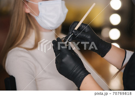 Close up of brow master hands in black gloves hold brush with henna. Background of customer in medical mask Close up of brow master hands in black gloves hold brush with henna. Background of customer in medical mask 70769818
