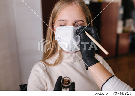Brow master in black protective mask applies henna on eyebrows for beautiful blond hair woman. Customer in medical mask. First person view Brow master in black protective mask applies henna on eyebrows for beautiful blond hair woman. Customer in medical mask. First person view 70769819