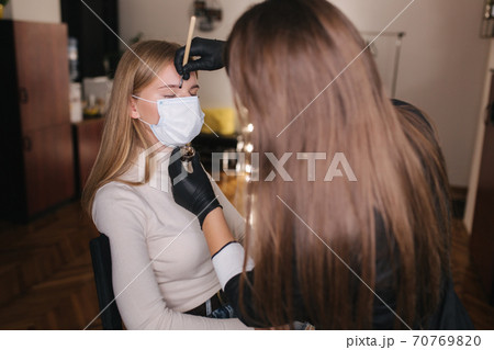 Brow master in black protective mask applies henna on eyebrows for beautiful blond hair woman. Customer in medical mask Brow master in black protective mask applies henna on eyebrows for beautiful blond hair woman. Customer in medical mask 70769820