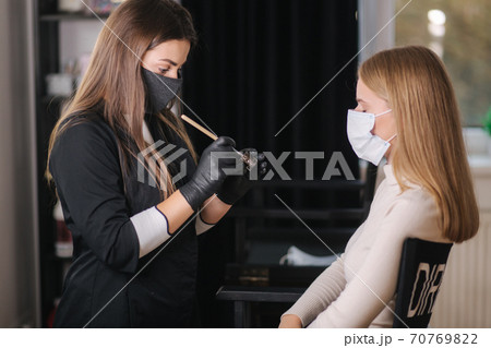 Brow master in black protective mask applies henna on eyebrows for beautiful blond hair woman. Customer in medical mask Brow master in black protective mask applies henna on eyebrows for beautiful blond hair woman. Customer in medical mask 70769822