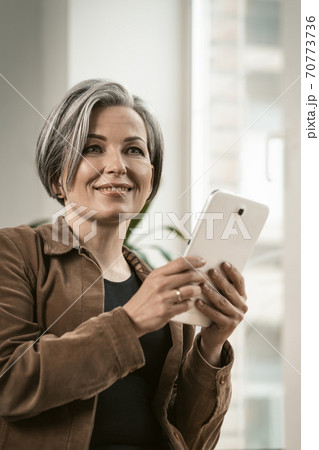 Pretty woman of retired age smiles communicating online on digital tablet while standing near window. Technology concept Pretty woman of retired age smiles communicating online on digital tablet while standing near window. Technology concept 70773736