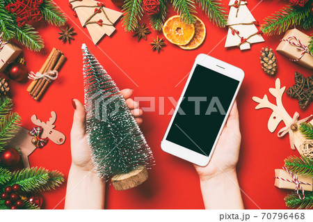 Top view of female hands holding phone in one hand and christmas tree in another hand on red background. New Year holiday concept. Mockup Top view of female hands holding phone in one hand and christmas tree in another hand on red background. New Year holiday concept. Mockup 70796468