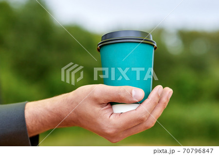 A man holds in his hand a paper cup with coffee or tea on a background of green foliage A man holds in his hand a paper cup with coffee or tea on a background of green foliage 70796847