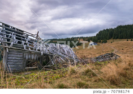 Old destroyed settlement in the mountains among the pastures with dried grass and fir-trees forest on the background. Old destroyed settlement in the mountains among the pastures with dried grass and fir-trees forest on the background. 70798174