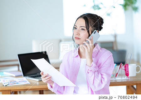 テレワーク リビングで書類を手にスマホで会話をする困った顔の若い女性 テレワーク リビングで書類を手にスマホで会話をする困った顔の若い女性 70802494