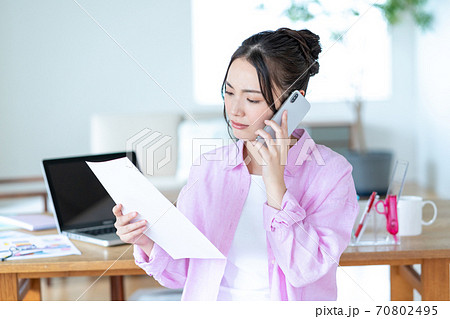 テレワーク  リビングで書類を手にスマホで会話をする困った顔の若い女性 70802495