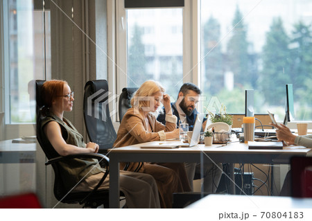 Happy mature businesswoman making notes by desk between two young colleagues Happy mature businesswoman making notes by desk between two young colleagues 70804183