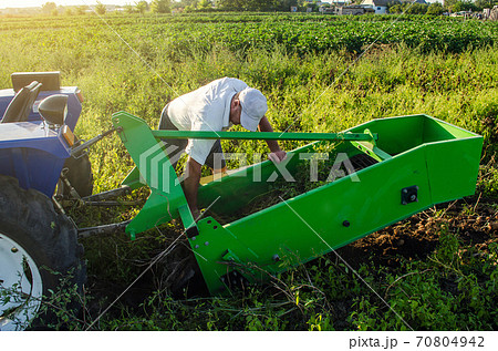A farmer examines a machine for digging out potato root vegetables. Exploitation and maintenance of equipment. Farming agriculture and farmland. Harvesting potatoes in autumn. 70804942