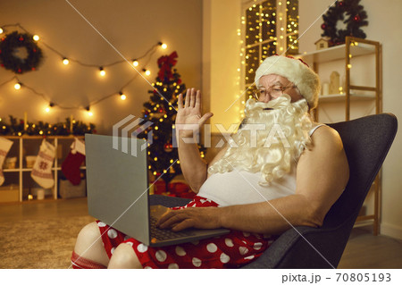 Santa sitting in armchair at home and waving hand at laptop screen starting online video call Santa sitting in armchair at home and waving hand at laptop screen starting online video call 70805193