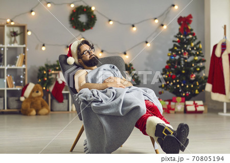 Tired young man dressed as Santa Claus sleeping peacefully in a cozy armchair under warm blanket Tired young man dressed as Santa Claus sleeping peacefully in a cozy armchair under warm blanket 70805194