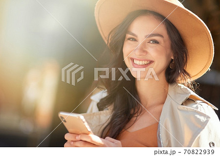 Portrait of a young beautiful and happy caucasian woman wearing hat using smartphone and smiling at camera while standing on the city street on a sunny day 70822939