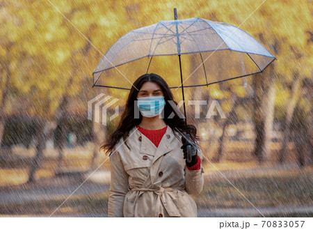 Beautiful young female in autumn coat standing in rainfall with umbrella and wearing antiviral mask 70833057