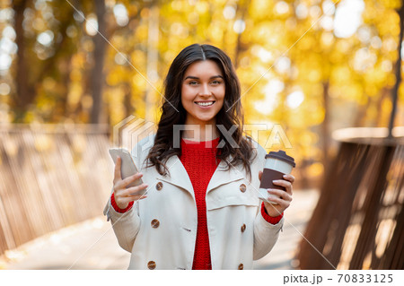 Portrait of pretty lady with coffee to go and smartphone smiling and looking at camera outdoors on cool autumn day 70833125