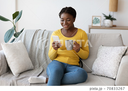 Domestic Relax. Positive black lady resting on couch with smartphone and coffee 70833267
