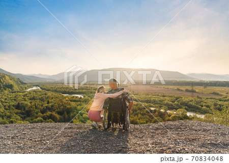 Beautiful woman hugs her disabled husband in wheelchair on a mountain hill at sunset. 70834048