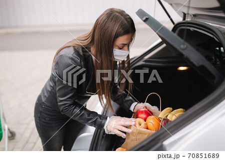A process of young attractive woman taking groceries from a supermarket from the trolley to car truck. Social distancing for protection, face mask, disposable gloves to prevent infection. Food A process of young attractive woman taking groceries from a supermarket from the trolley to car truck. Social distancing for protection, face mask, disposable gloves to prevent infection. Food 70851689