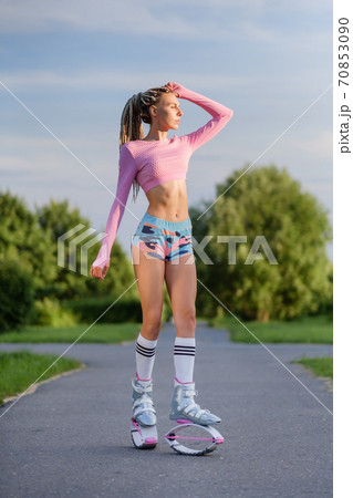 A young slender beautiful woman with dreadlocks in sportswear trains in the park outdoors. Kangoo jumps training. 70853090