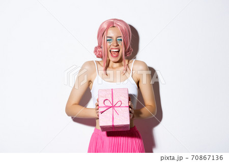Cheerful birthday girl looking excited, wearing pink wig, shouting of joy, receiving bday gift, standing over white background and celebrating 70867136