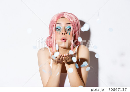 Close-up of lovely pretty party girl with pink wig and bright makeup, blowing confetti from palm, celebrating holiday, standing over white background 70867137
