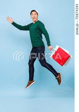 Happy young handsome Asian man wearing Christmas hat jumping with shopping bags 70874284