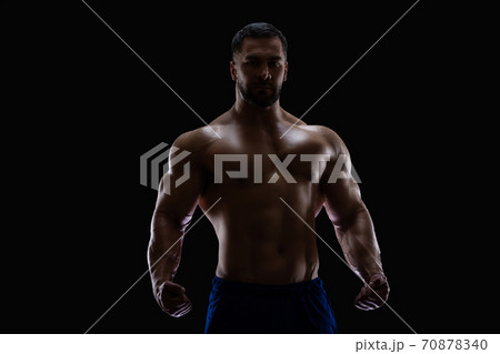 Portrait of a bodybuilder standing isolated on black background in a shadow with clenched fists to show off his muscles Portrait of a bodybuilder standing isolated on black background in a shadow with clenched fists to show off his muscles 70878340