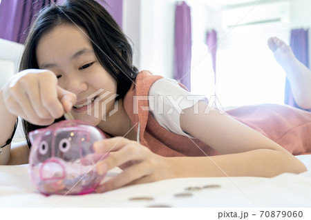 Happy smiling asian child girl put coin into piggy bank while lying on her bed,student saving money for the future in education,health care,medical insurance,concept of financial planning,lifestyle 70879060