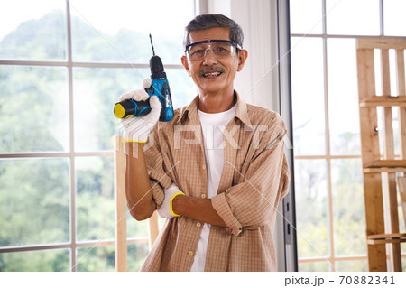 Asian senior carpenter safety goggles hold electric drill in carpentry workshop. Concept senior people doing hobby after retirement. Asian senior carpenter safety goggles hold electric drill in carpentry workshop. Concept senior people doing hobby after retirement. 70882341