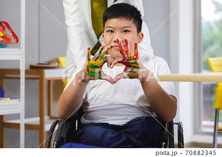 Disabled boy on wheelchair painting his hand with yellow, red and green in art class. Concept children learning in school. Disabled boy on wheelchair painting his hand with yellow, red and green in art class. Concept children learning in school. 70882345