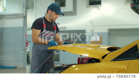 Professional car mechanic is working on a vehicle in a service. Happy repairman is closing the hood. Man Looks at a Camera and Smiles. Professional car mechanic is working on a vehicle in a service. Happy repairman is closing the hood. Man Looks at a Camera and Smiles. 70890778