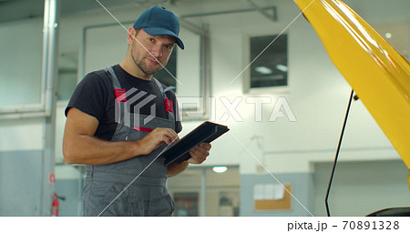 In a garage a mechanic using the tablet makes the oil and engine check to the car smiles. Concept of: security, safety, insurance and professionalism, revisions, technology. 70891328