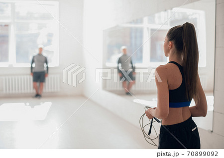 In a bright gym with large windows a young female athlete jumps on a rope under the supervision of a coach 70899092
