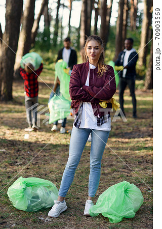 Active pretty girl with full rubbish bags in backdrop of her friends volunteers which are picking up rubbish at forest. Active pretty girl with full rubbish bags in backdrop of her friends volunteers which are picking up rubbish at forest. 70903569