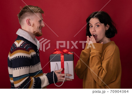 Happy man presents Christmas gift box to surprised girlfriend on red background Happy man presents Christmas gift box to surprised girlfriend on red background 70904364