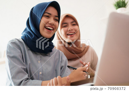 attractive cheerful young muslim business woman working on laptop and smiling while sitting at her desk modern office with her friend 70908372
