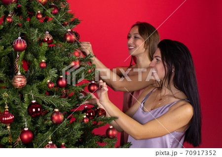Happy lesbian couple joyfully decorate Christmas tree together on red backdrop 70917352