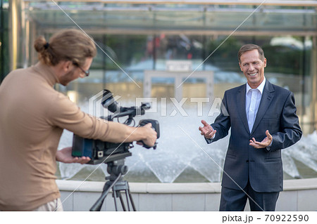 Fair-haired operator filming mature male reporter standing in front of fountain Fair-haired operator filming mature male reporter standing in front of fountain 70922590