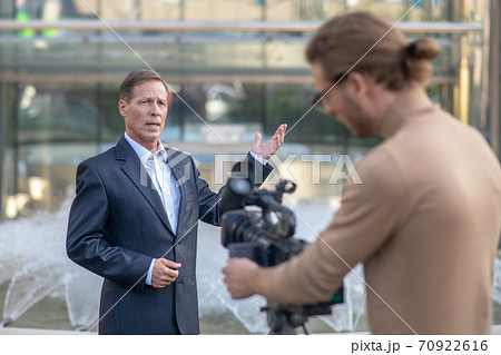 Long-haired operator filming mature male reporter in front of fountain 70922616