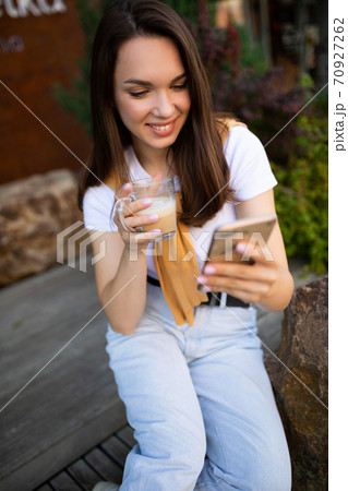charming free young woman in casual look is drinking coffee while sitting in a summer park charming free young woman in casual look is drinking coffee while sitting in a summer park 70927262