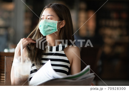 Young attractive woman wearing surgical mask sitting alone at the public area in natural sunlight atmosphere, New normal concept for everyone wearing mask in public place, social distancing lifestyle. 70928300